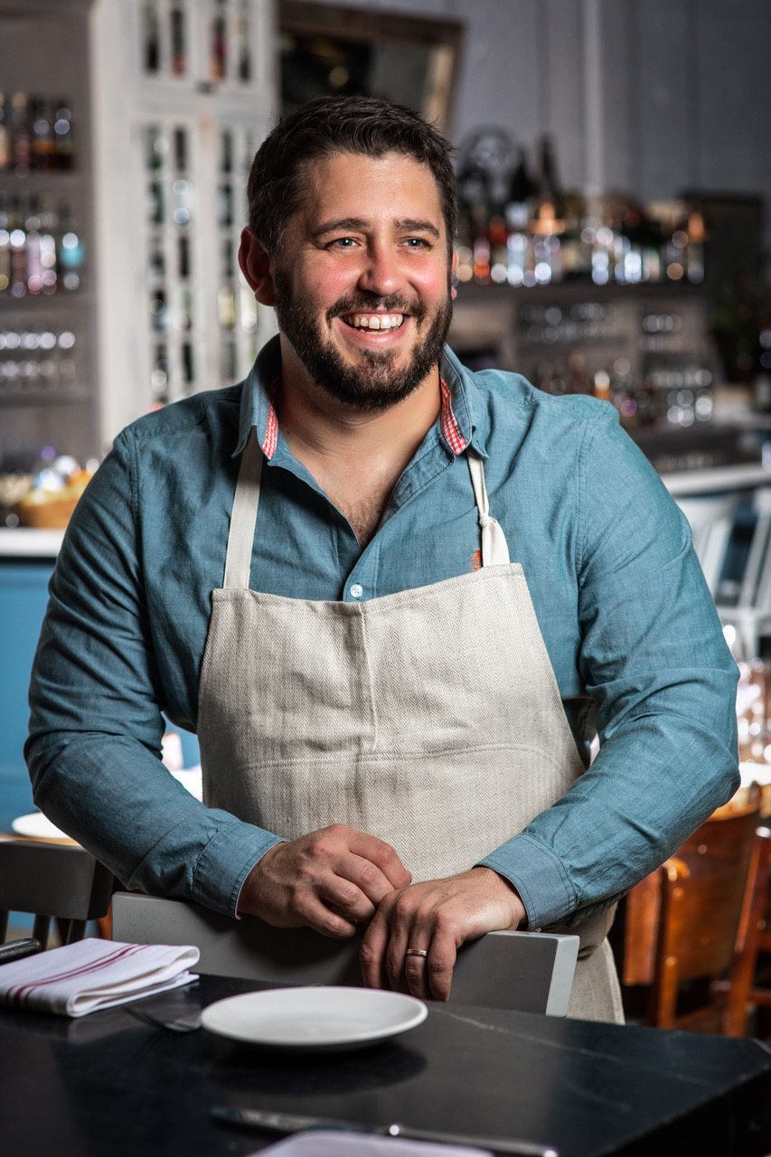 Chef Will Gilson in an apron, leaning on a chair in the dining room